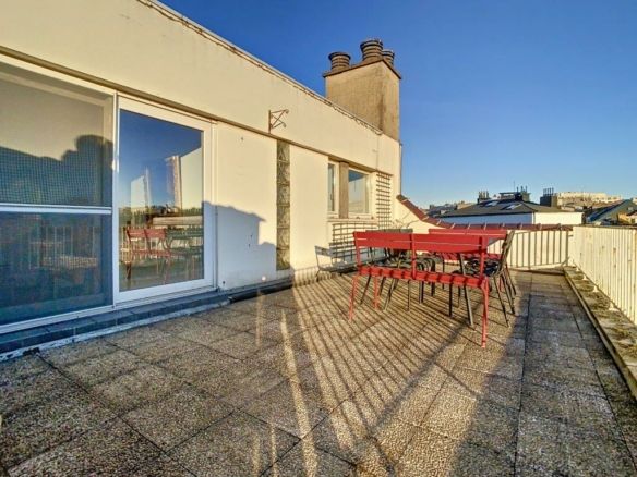 Terrasse urbaine ensoleillée avec mobilier rouge, vue dégagée, grande porte vitrée, ambiance contemporaine.