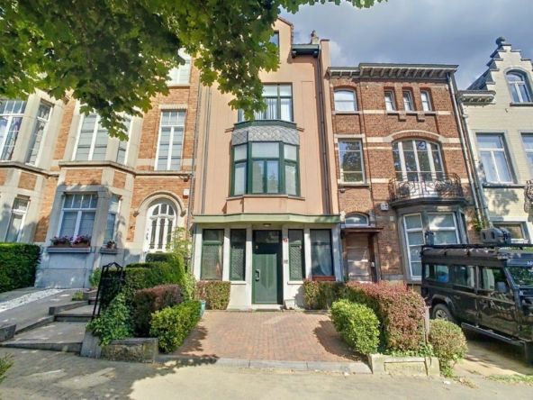 Stadsstraat met bakstenen gebouwen, oldtimers, goed onderhouden tuinen en een bewolkte lucht.