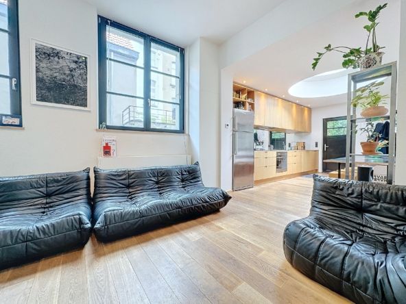 Lichte woonkamer met zwarte banken, moderne open keuken, planten en grote houten ramen.