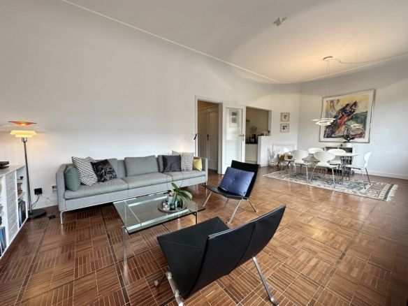 Salon moderne lumineux avec parquet, canapé gris, mobilier transparent et œuvre d’art colorée.