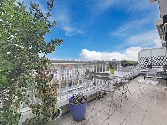 Balcon lumineux avec vue urbaine, ciel bleu, meubles en métal et plantes colorées.