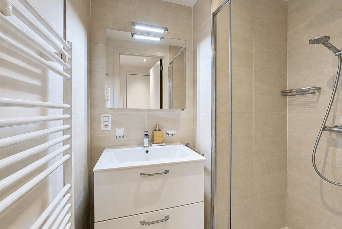 Modern beige-tiled bathroom with a white vanity, mirror, and glass shower enclosure.