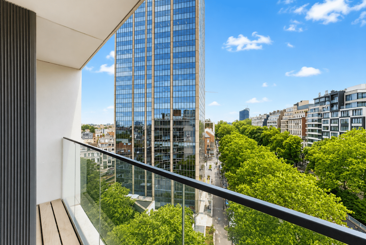 Balcony with glass railing overlooking a tree-lined street and a tall glass skyscraper against a blue sky