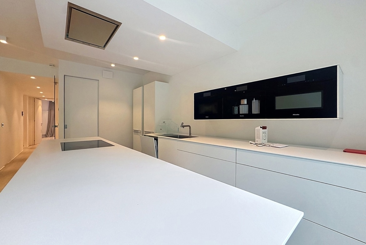 Cuisine équipée moderne blanche avec îlot, électroménagers encastrés et couloir d’appartement.