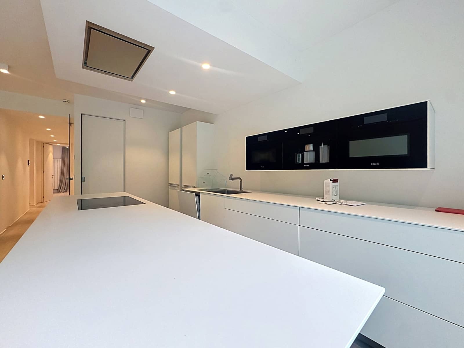 Cuisine Équipée Moderne – You Real Estate Agency Cuisine équipée moderne blanche avec îlot, électroménagers encastrés et couloir d’appartement.