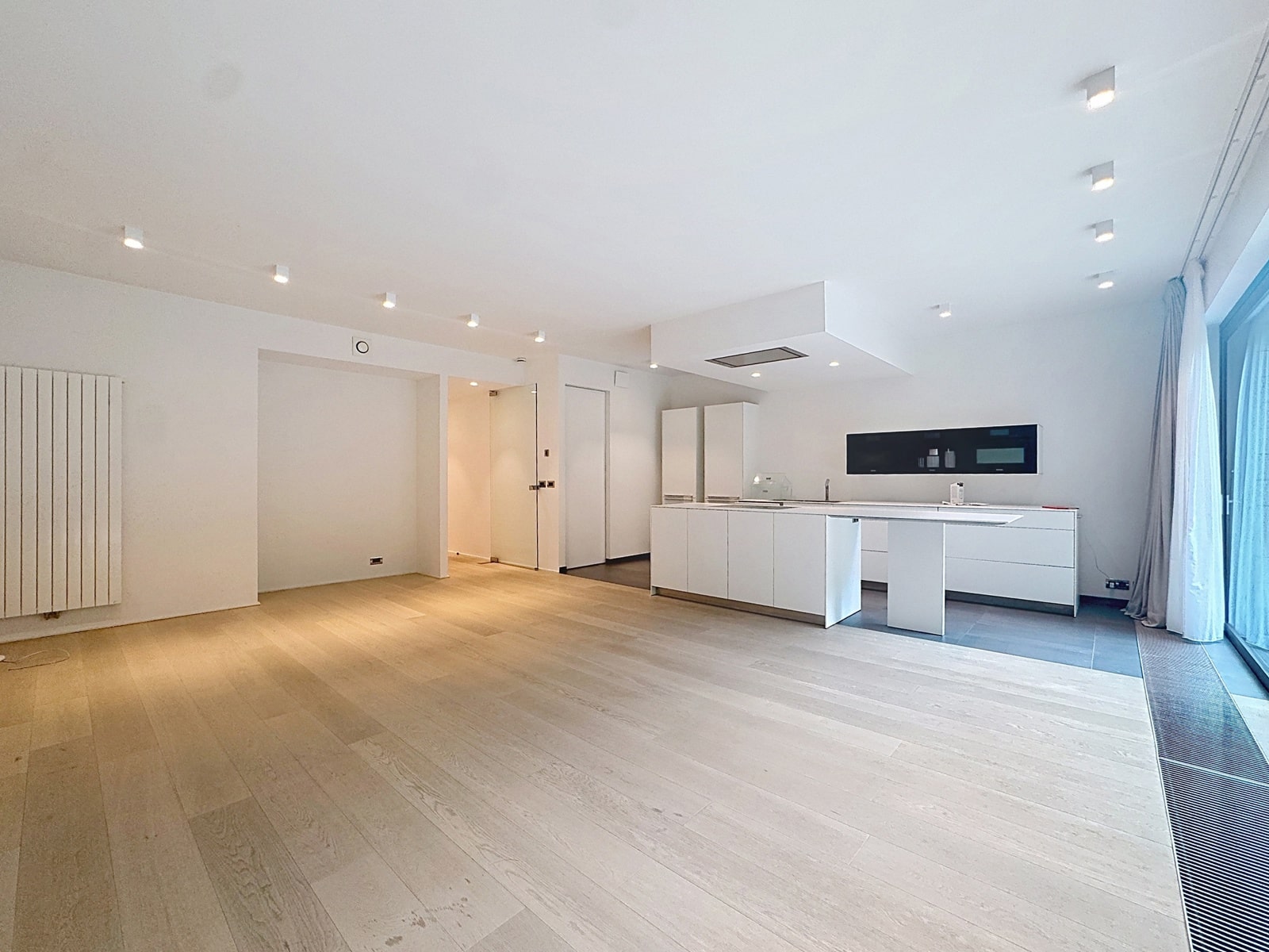 Séjour Ouvert avec Cuisine Moderne – You Real Estate Agency Open-plan kitchen with white cabinets and island, large windows with sheer curtains, light wood floors menacing natural lighted space