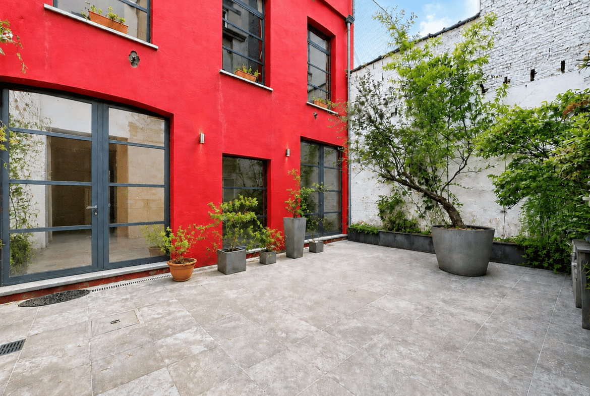 Red-painted building courtyard with large glass doors, concrete tiles, and a variety of potted plants along the wall.