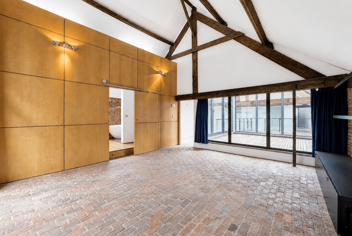 Grand salon loft avec plafond voûté, poutres apparentes, briques et terrasse vitrée.
