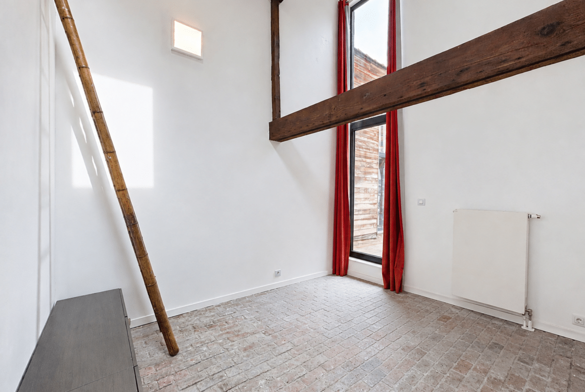 Pièce lumineuse d’appartement avec poutres apparentes, échelle en bambou et grande fenêtre.
