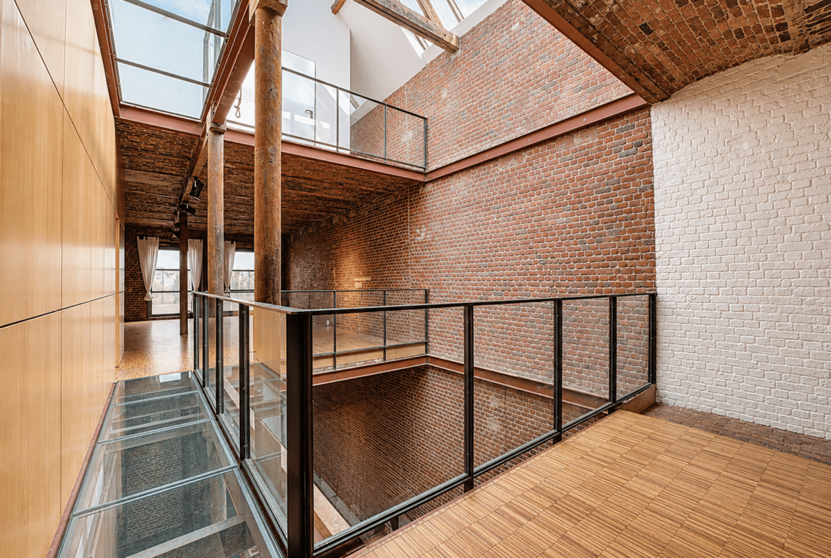 Atrium lumineux d’un loft rénové avec murs en briques, verrière et passerelles vitrées.