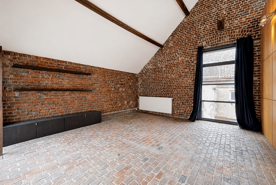 Brick-walled loft interior with exposed wooden beams, low dark cabinets along the left wall, and a large window with dark curtains.