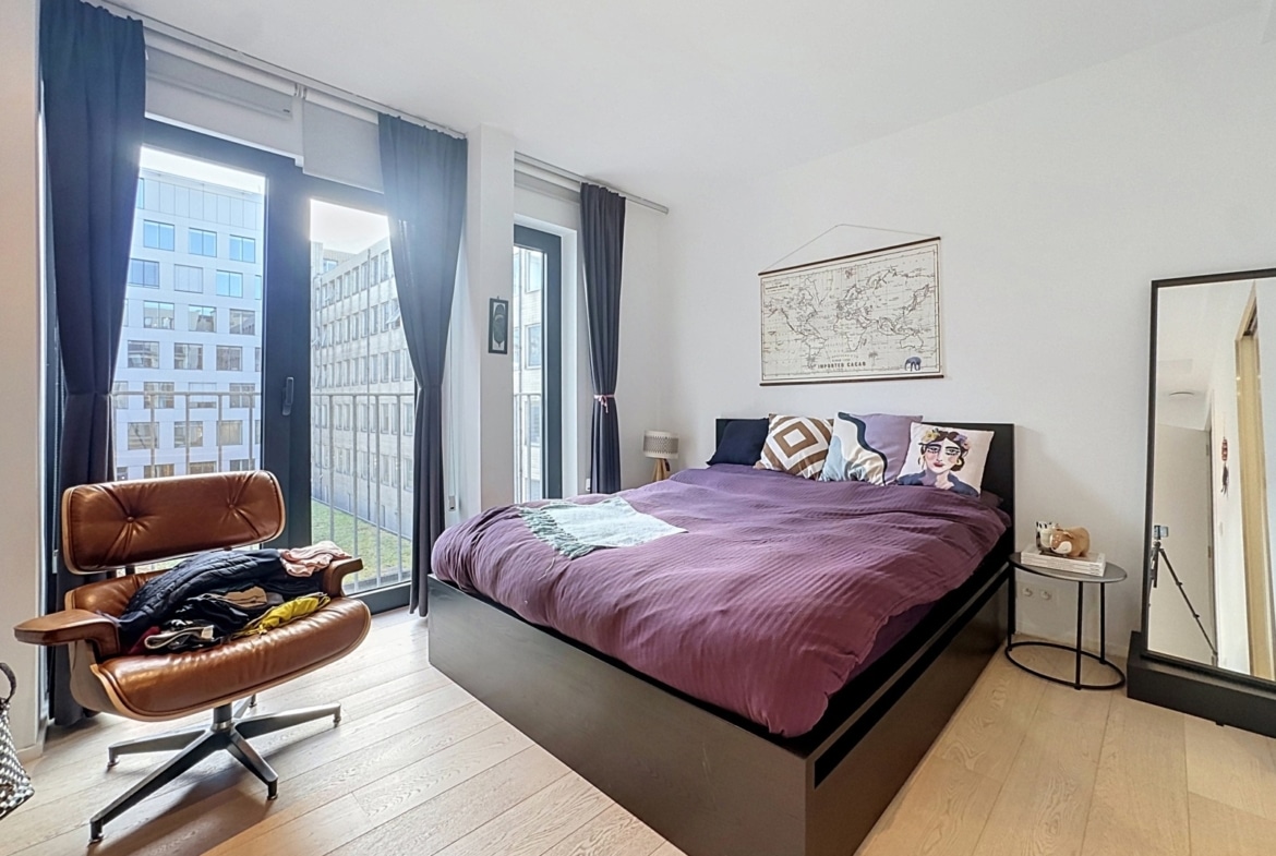 A modern bedroom with a purple duvet on the bed against a white wall, tall windows with dark curtains, and a brown leather chair by the window