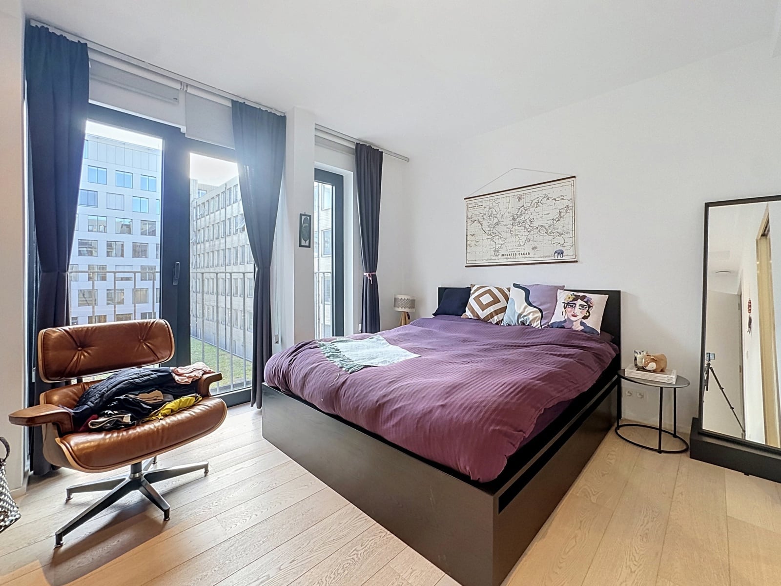 whise photo 11 A modern bedroom with a purple duvet on the bed against a white wall, tall windows with dark curtains, and a brown leather chair by the window