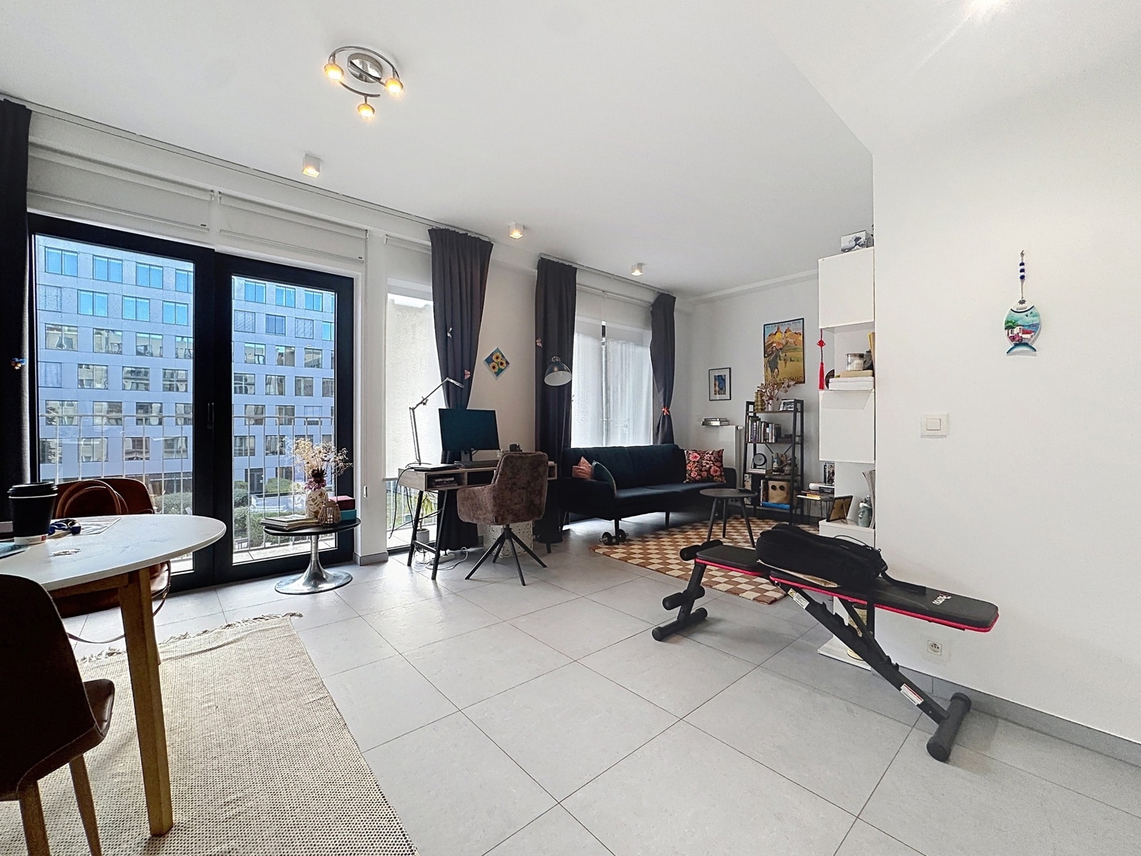 whise photo 6 A bright living space with large glass doors offering a view of the city, a study desk, and a dark sofa set next to a gym bench in the foreground.
