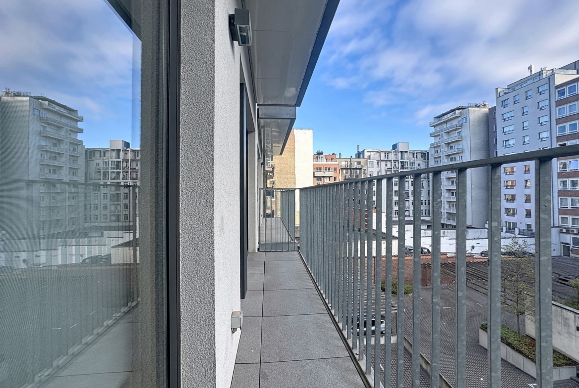 A narrow apartment balcony with a metal railing, gray tiles, and tall city buildings in the distance beneath a blue sky.