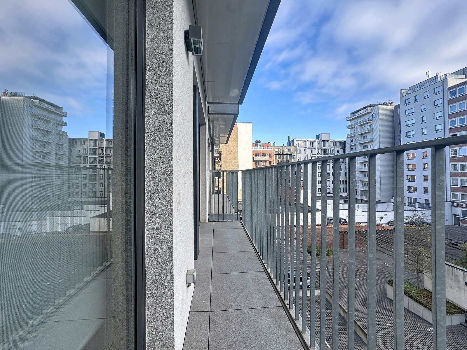 whise photo 10 A narrow apartment balcony with a metal railing, gray tiles, and tall city buildings in the distance beneath a blue sky.