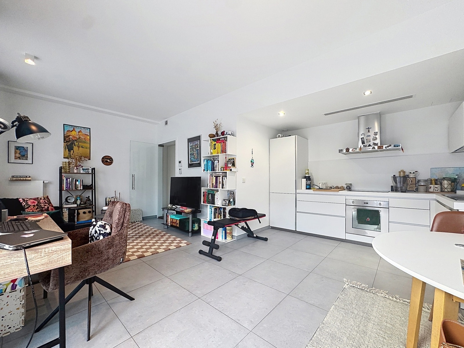Bright Living Room with Open Kitchen – You Real Estate Agency Bright, open-concept living area and white kitchen with a sofa, bookshelf, and exercise bench.