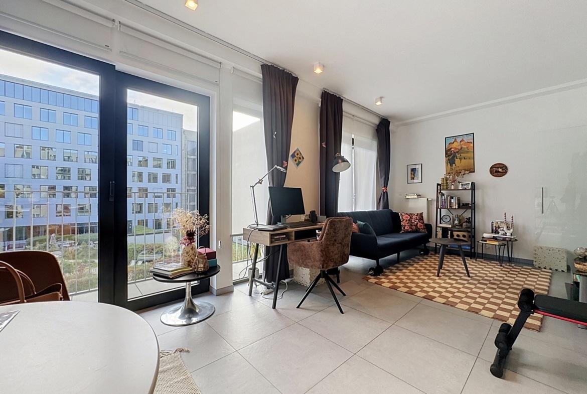 A bright living room with large glass doors leading to a balcony, a home office set up by the window, and a cozy seating area.