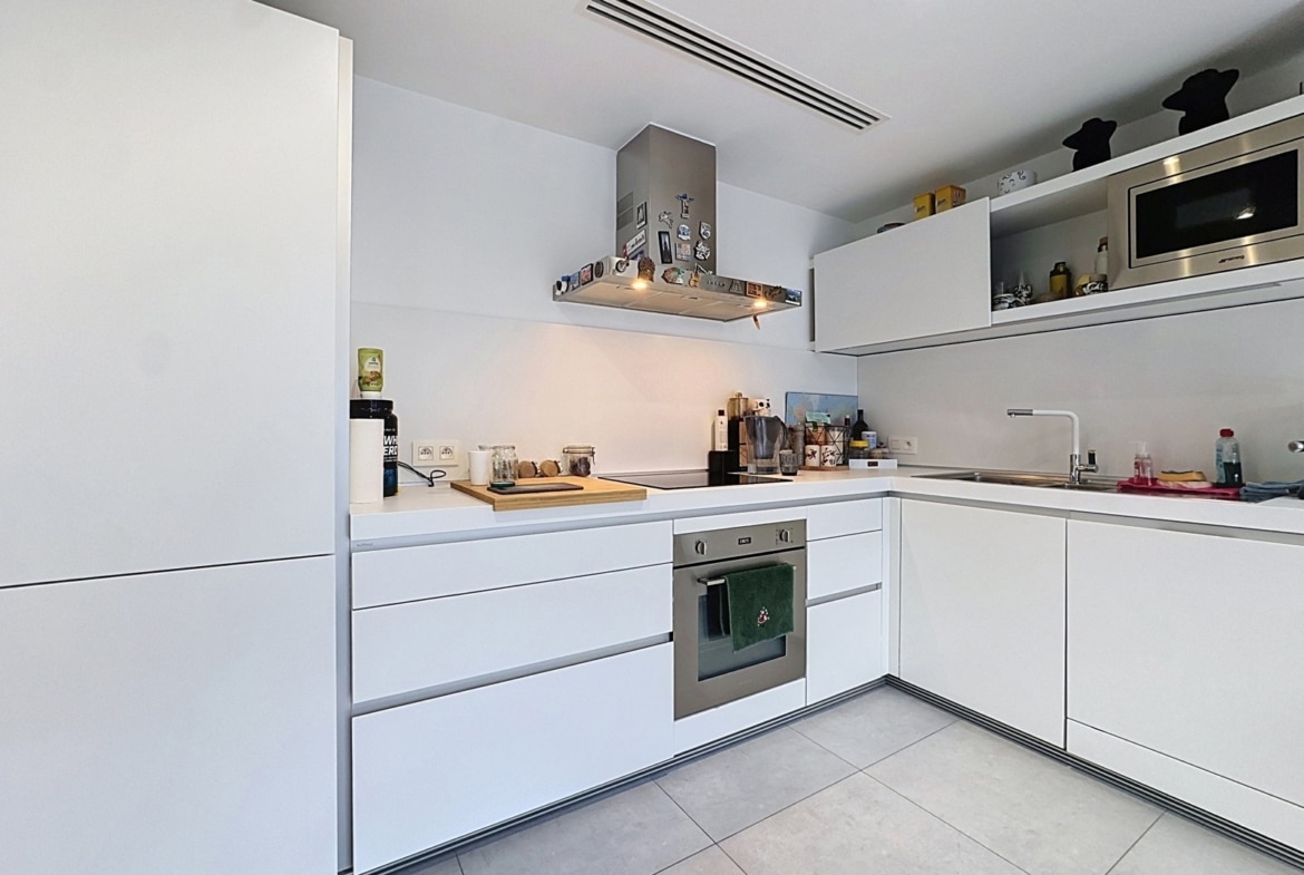A contemporary white kitchen with a built-in oven, a range hood covered in magnets, and white cabinets along an L-shaped counter.
