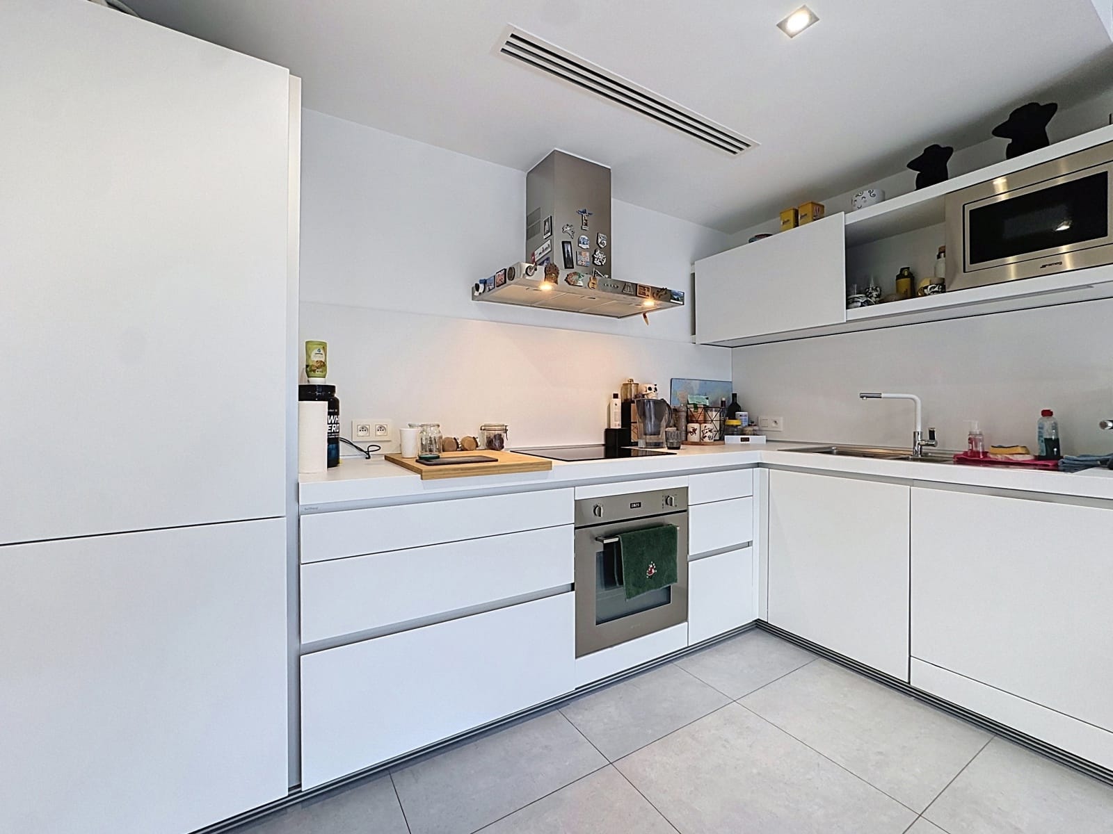 whise photo 3 A contemporary white kitchen with a built-in oven, a range hood covered in magnets, and white cabinets along an L-shaped counter.