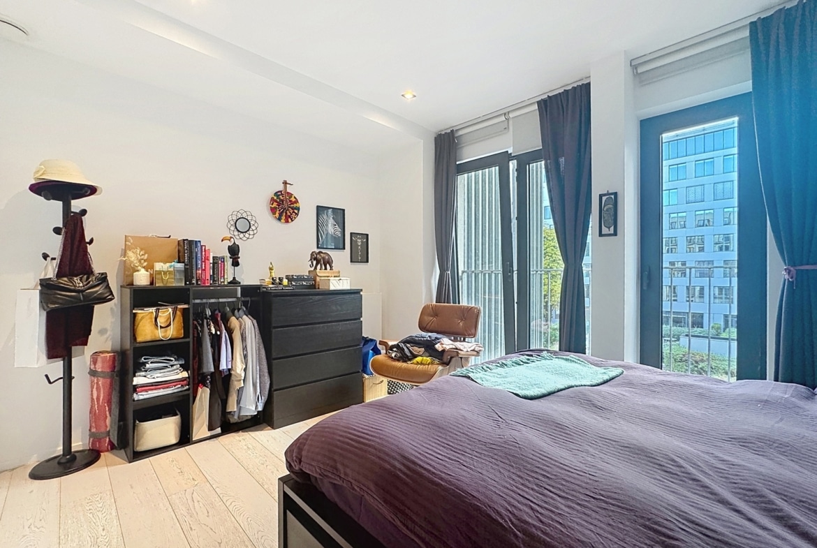 A bright bedroom in a modern apartment with a double bed, storage space, and large picture windows.