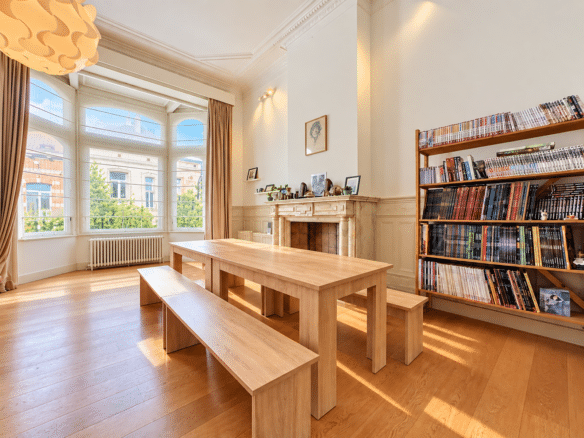 Bright sunlit library reading room with a large bay window a long wooden table and benches and a wall of magazine filled shelves to the right   appartement 1 chambre  saint gilles