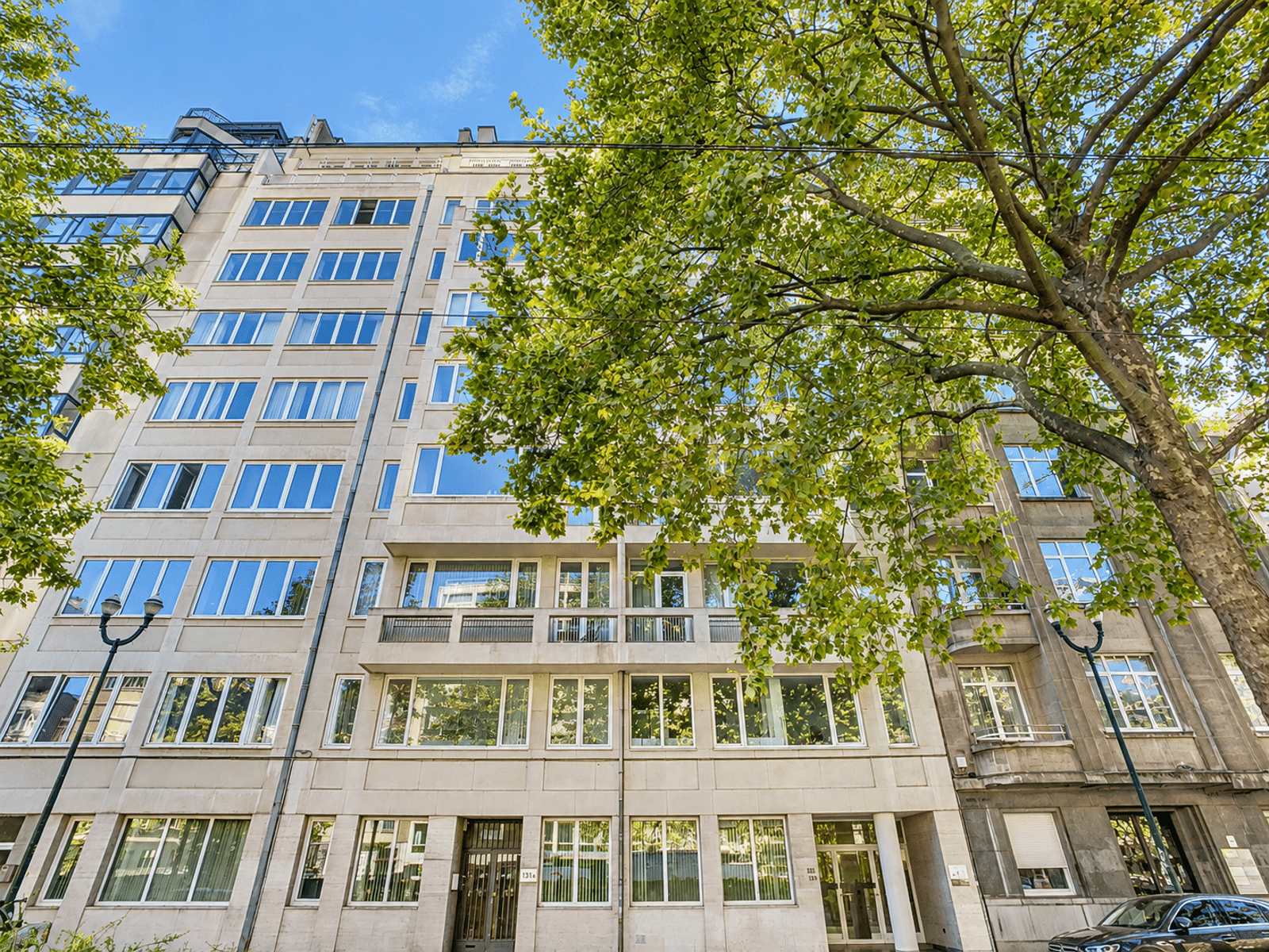 Exterior View of a Modern Building – You Real Estate Agency A modern residential building with apartments, a light-colored facade, and trees lining the street.
