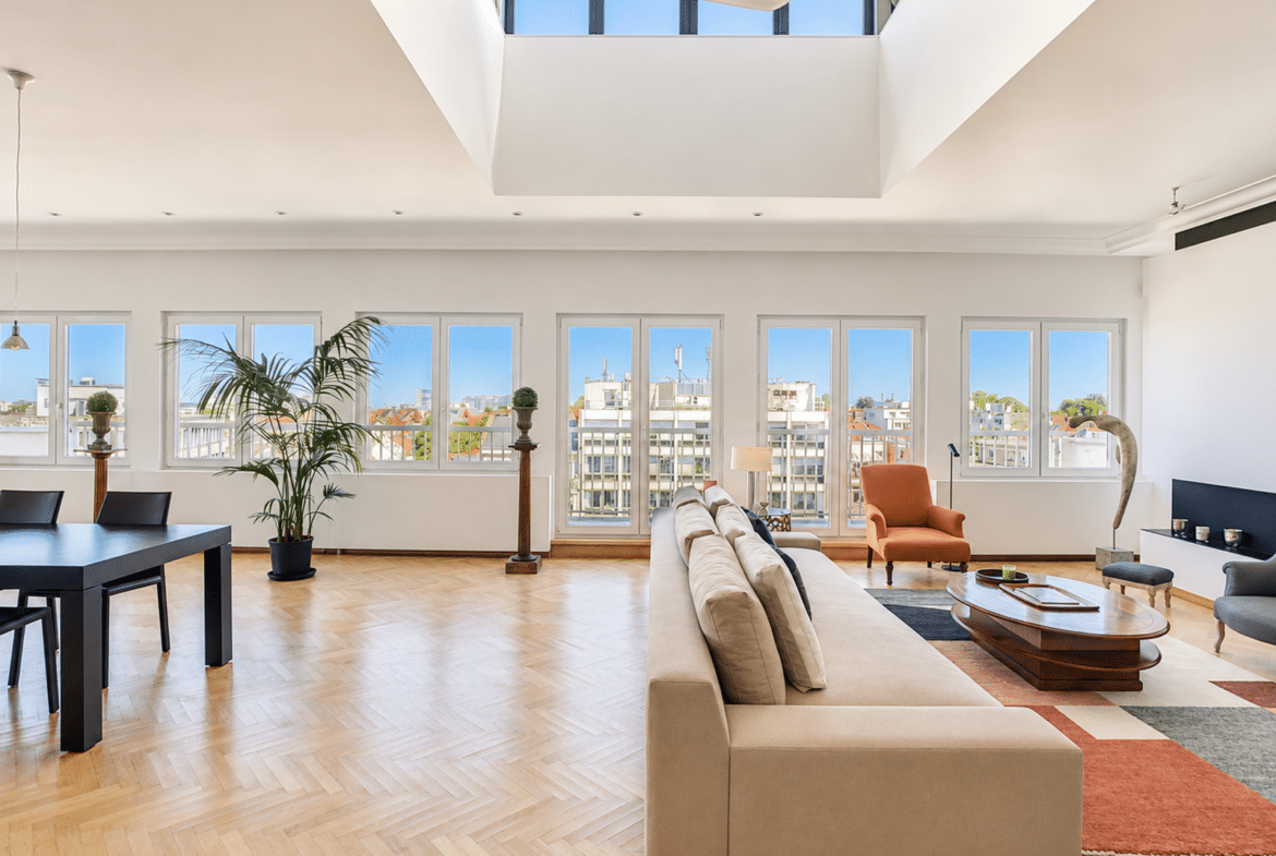 A bright, open living room with a beige sectional, an orange armchair, and a wooden coffee table; large windows look out onto the city skyline.