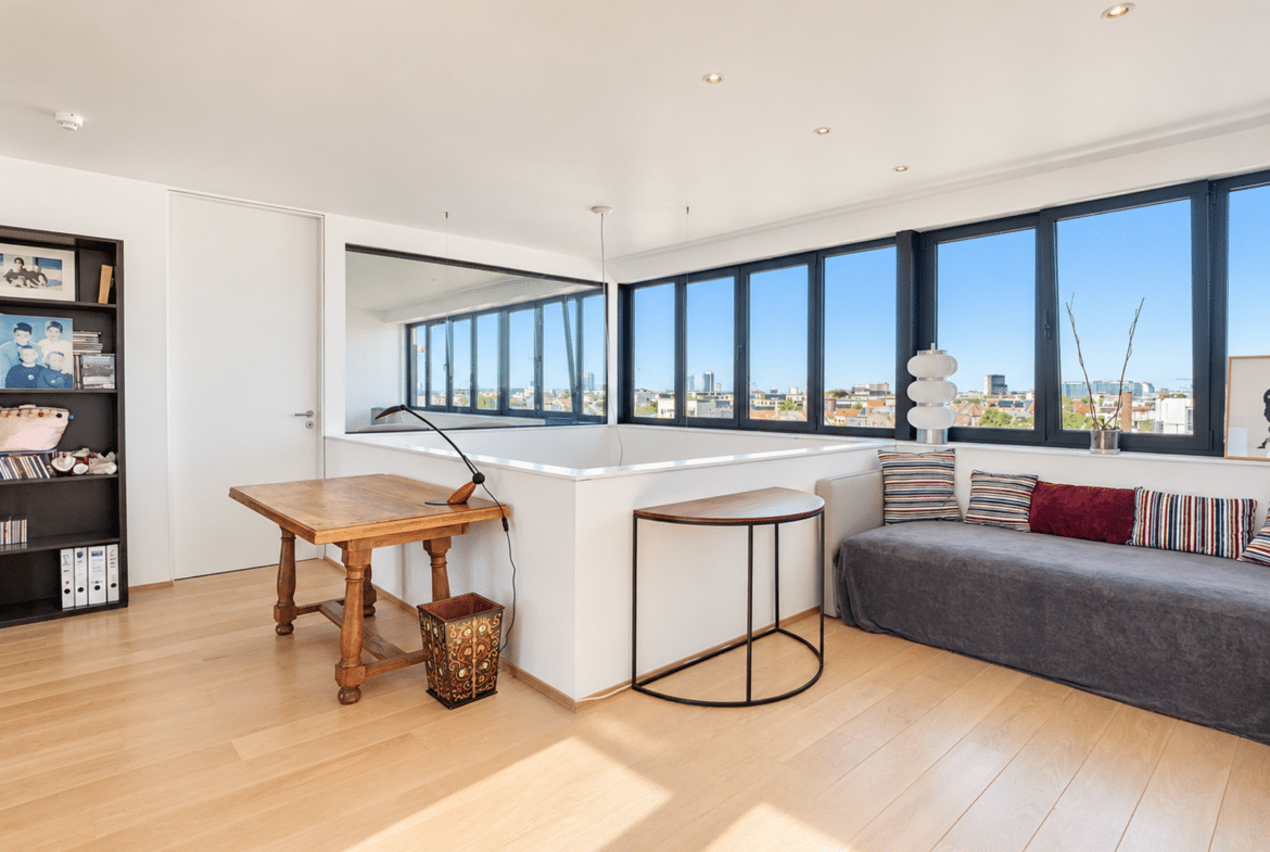 A bright living room in a modern apartment with large windows, light-colored hardwood floors, and a city view.