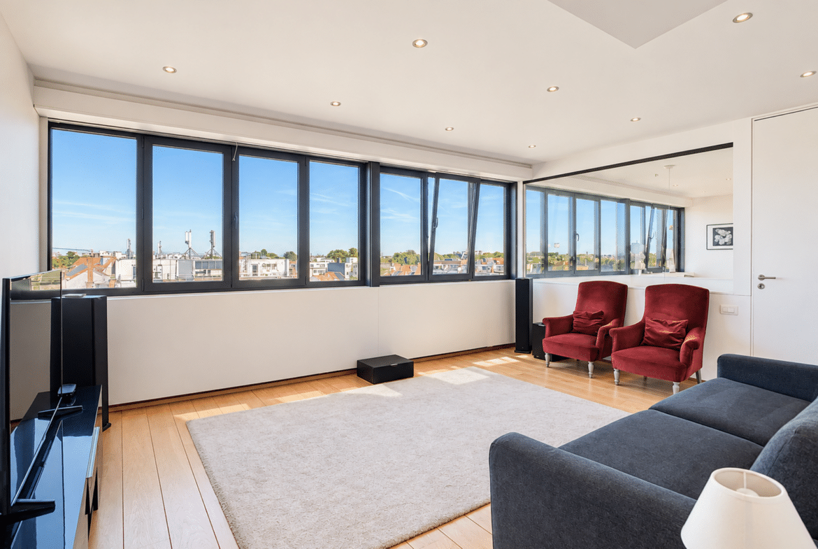 A bright living room in a modern apartment with large windows, light-colored hardwood floors, and red armchairs.