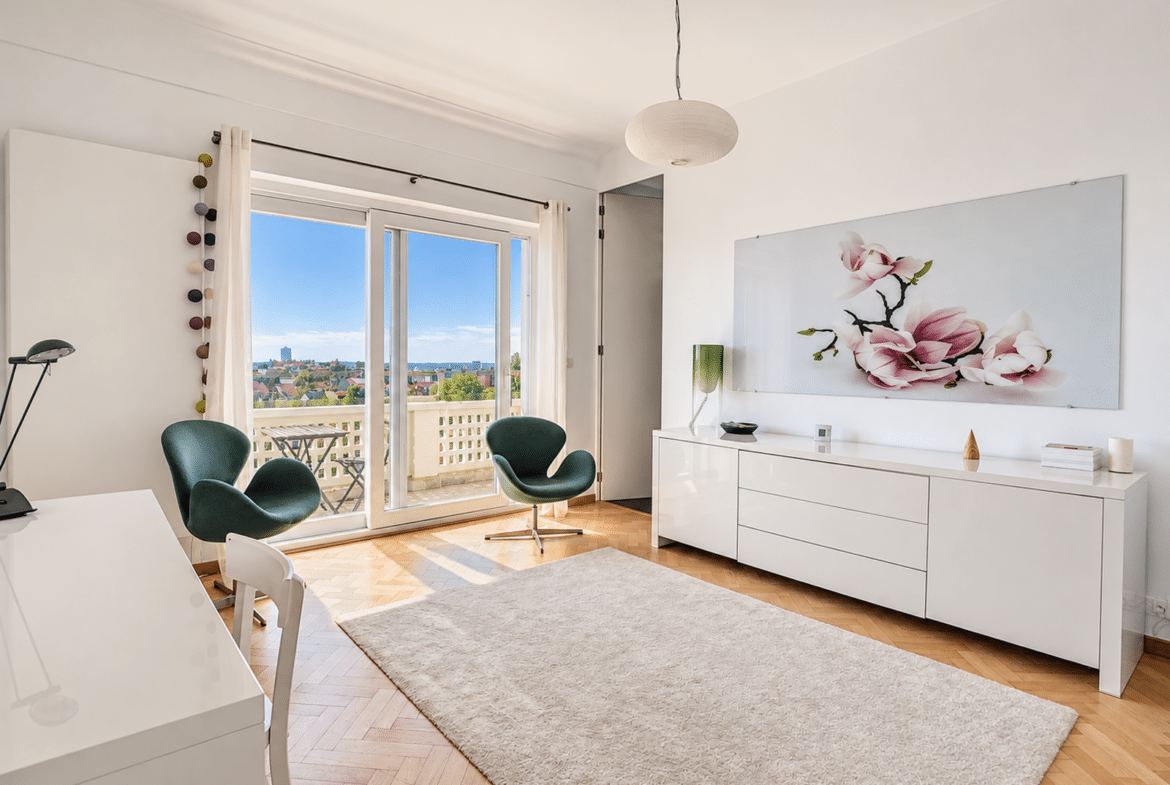 A bright, minimalist living room with white furniture, two green lounge chairs, a cream-colored rug, and balcony doors offering a view of the city.
