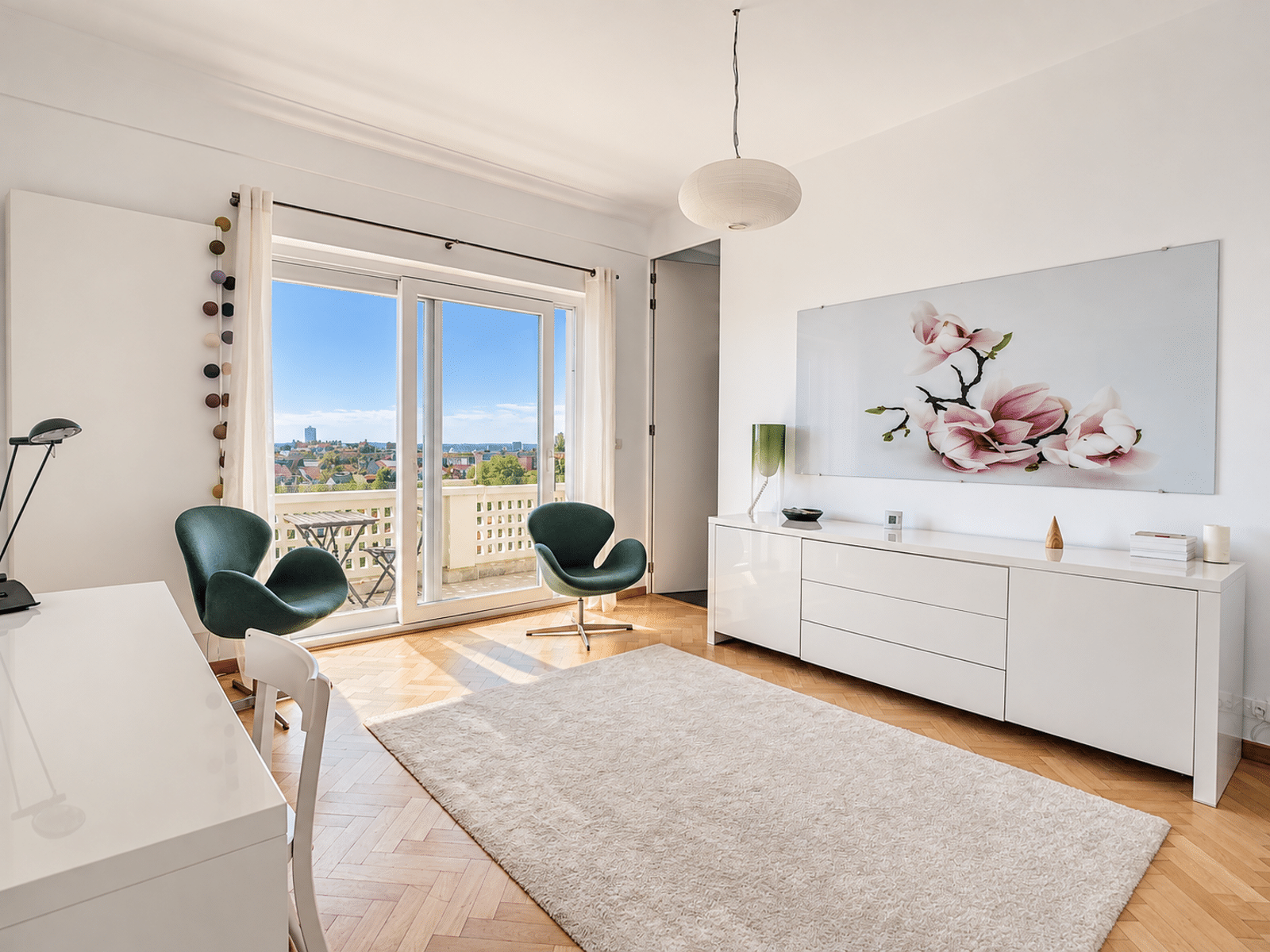Bright living room with balcony - You Real Estate Agency A bright, minimalist living room with white furniture, two green lounge chairs, a cream-colored rug, and balcony doors offering a view of the city.