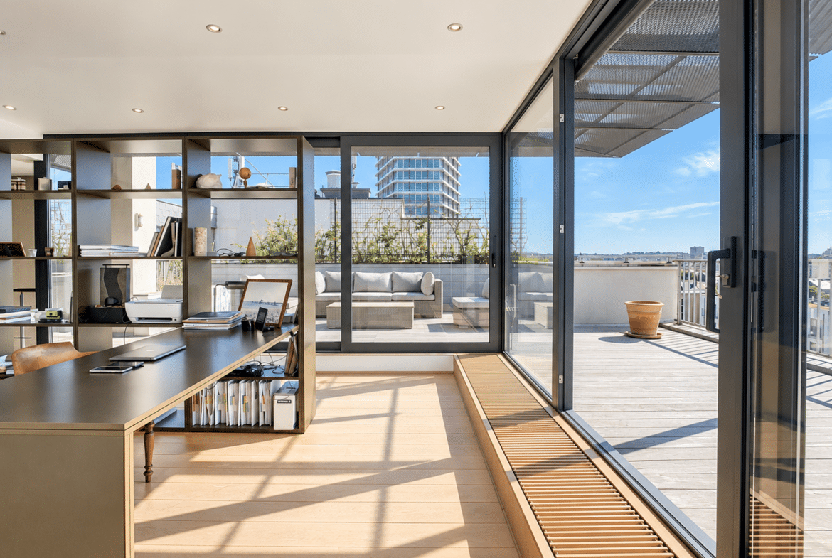 A bright, open-plan office with a long, dark desk, built-in shelves, and large glass doors opening onto a sunlit outdoor deck.