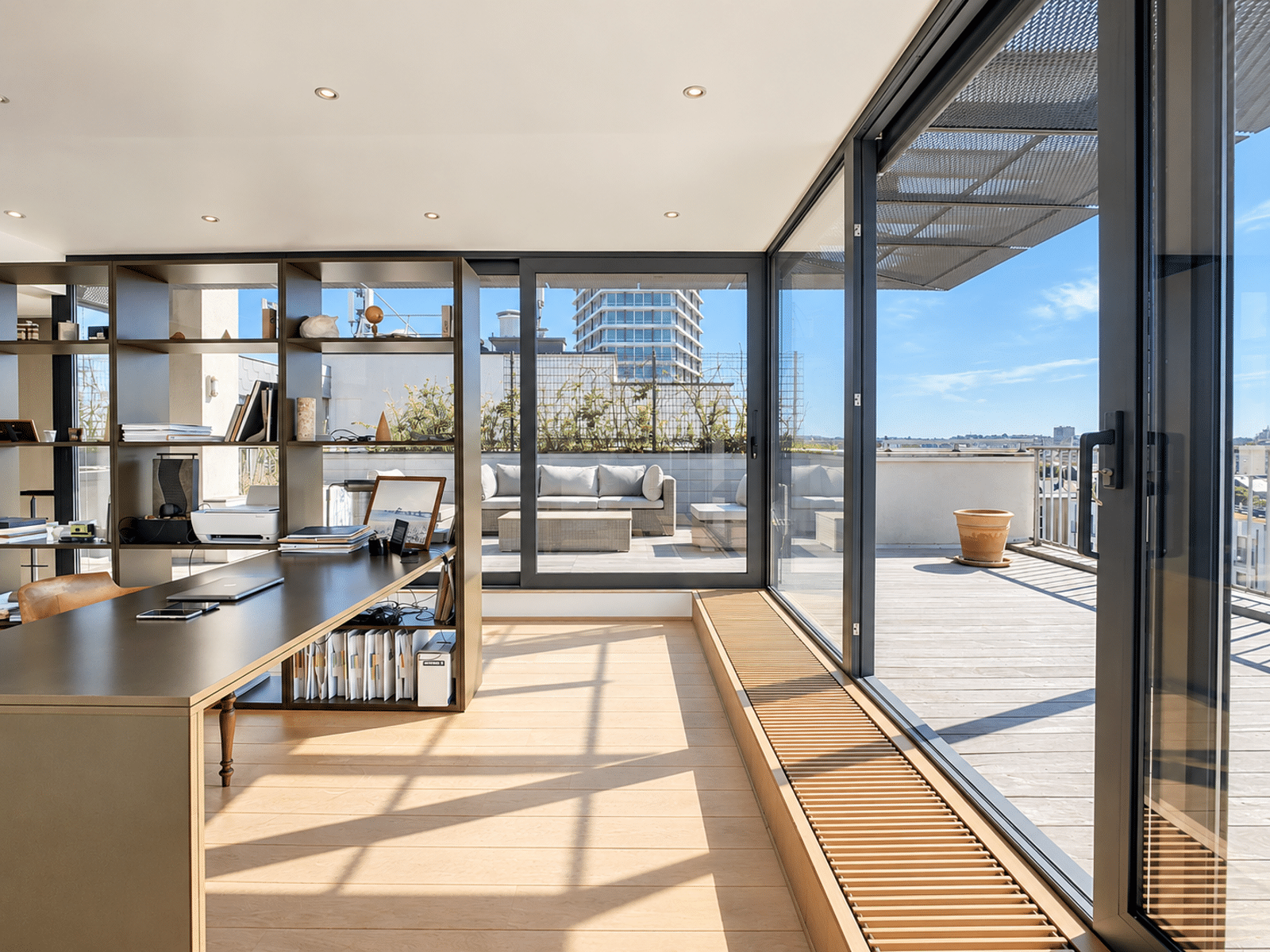 whise photo 2 A bright, open-plan office with a long, dark desk, built-in shelves, and large glass doors opening onto a sunlit outdoor deck.