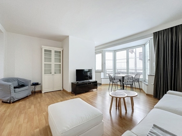 Bright living room with white furniture a tv on a black stand a glass front cabinet and a bay window with a round dining table and wire chairs   appartement 1 chambre  bruxelles