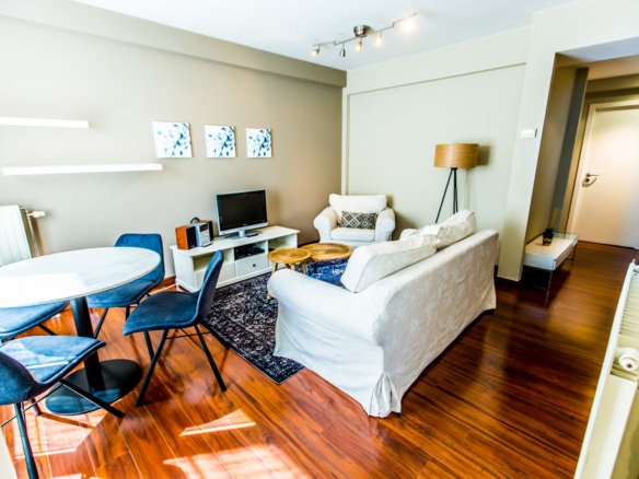 Bright living room with a white sofa armchairs round dining table with blue chairs and a tv on a white stand Three blue tinted wall art panels on beige wall wood floor   appartement 1 chambre  bruxelles