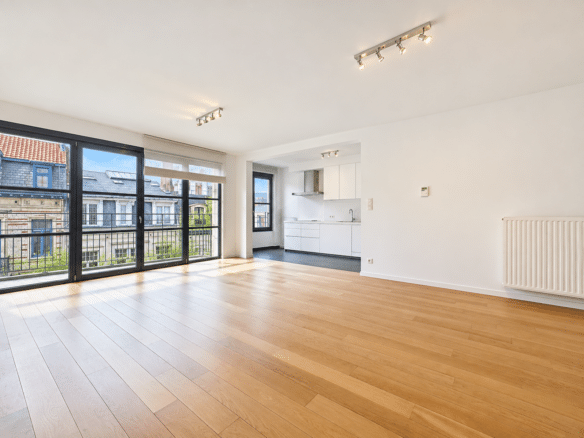 Open plan living room with light wood flooring white walls and large black framed windows letting in daylight opening to a balconystreetscape outside A white kitchen with integrated cabinets is visible in the back right corner   appartement 3 chambres  ixelles