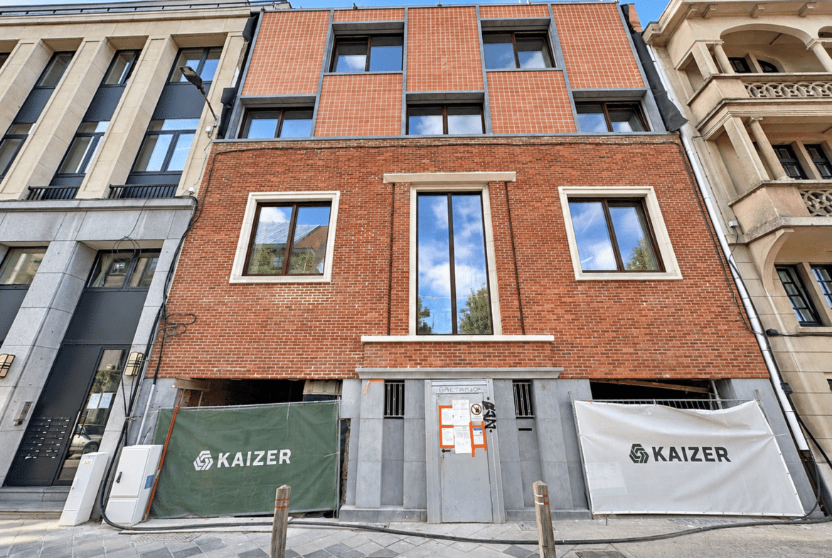 Een bakstenen gebouw in renovatie met een geelrode bakstenen gevel en grote ramen; bouwhekken met KAIZER-logo’s op de begane grond.