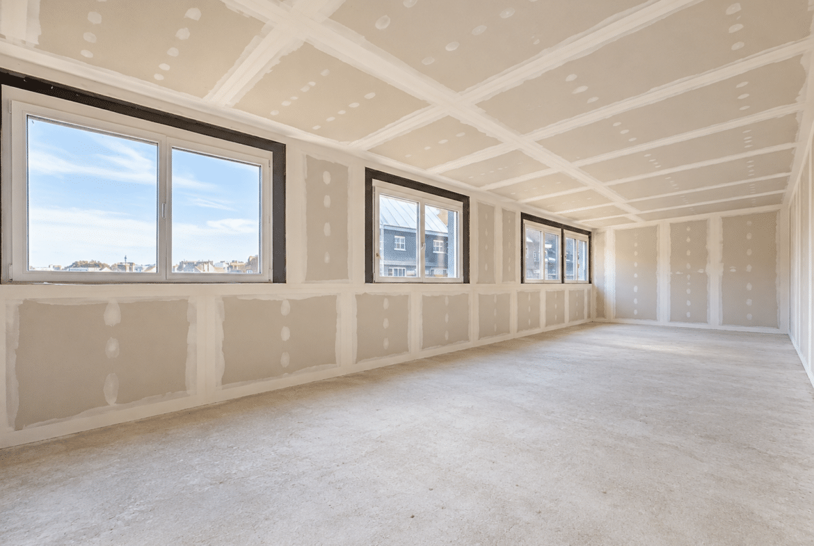 Grote woonkamer in een appartement dat momenteel wordt gerenoveerd, met grote ramen en gipsplaatwanden.