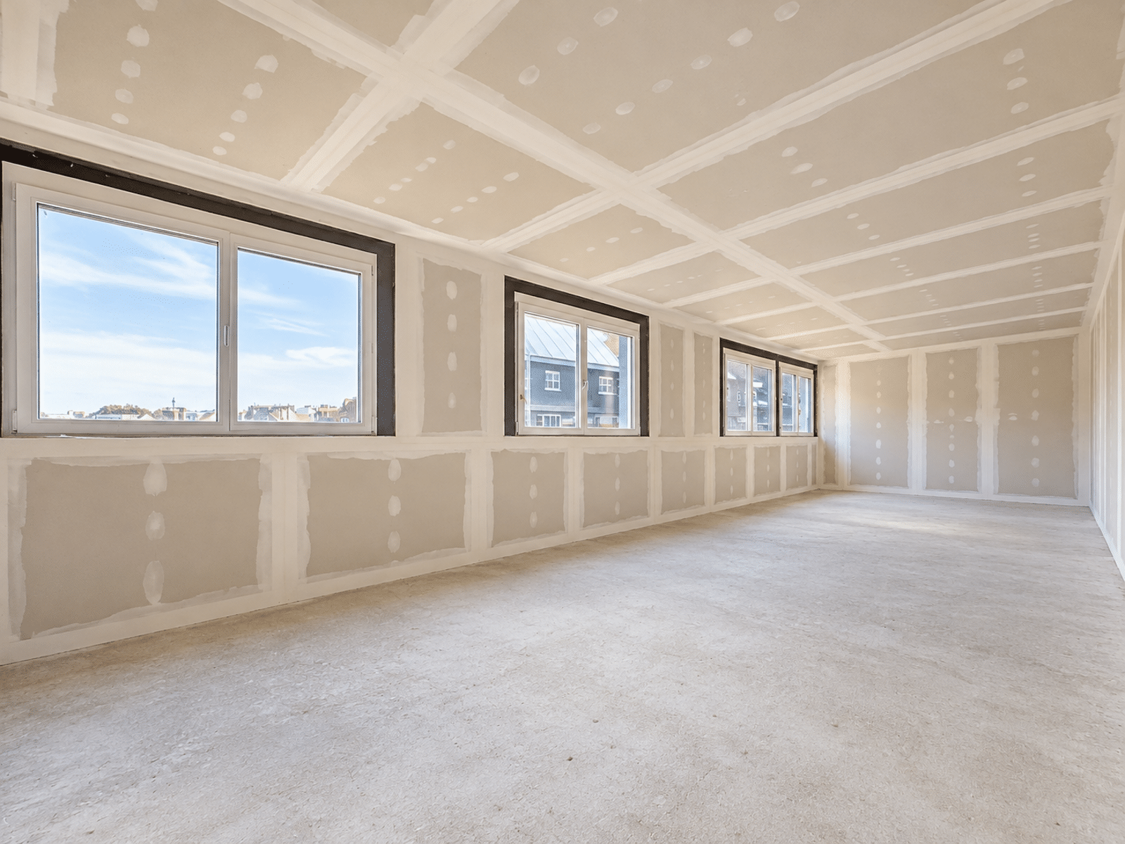 Lichte woonkamer in renovatie – You Real Estate Agency Grote woonkamer in een appartement dat momenteel wordt gerenoveerd, met grote ramen en gipsplaatwanden.