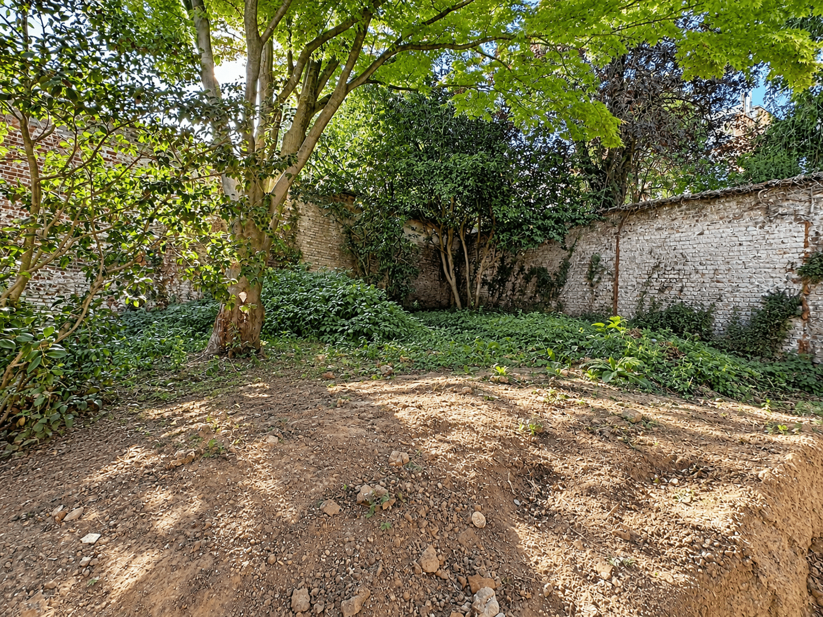 Tuin met bomen en omheind terrein – You Real Estate Agency Een omheinde tuin met bomen bij een huis met bakstenen muren.
