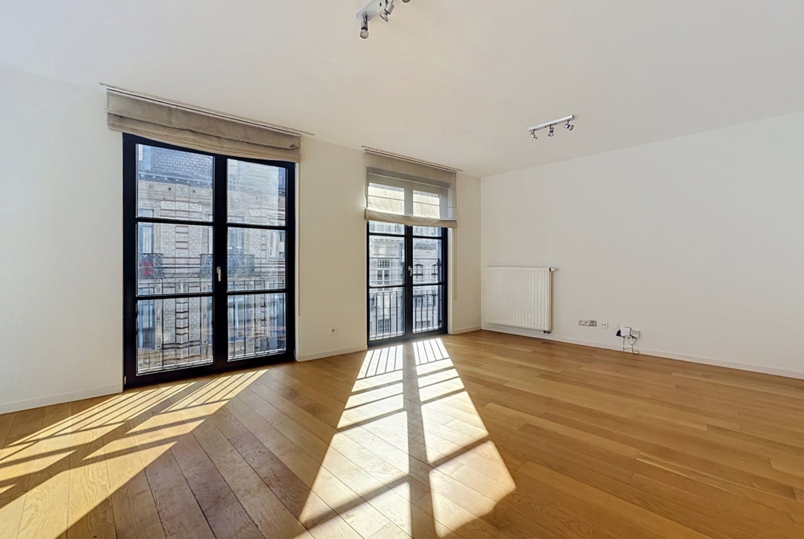 Lichte woonkamer in een modern appartement met parketvloer, grote ramen en zonwering.