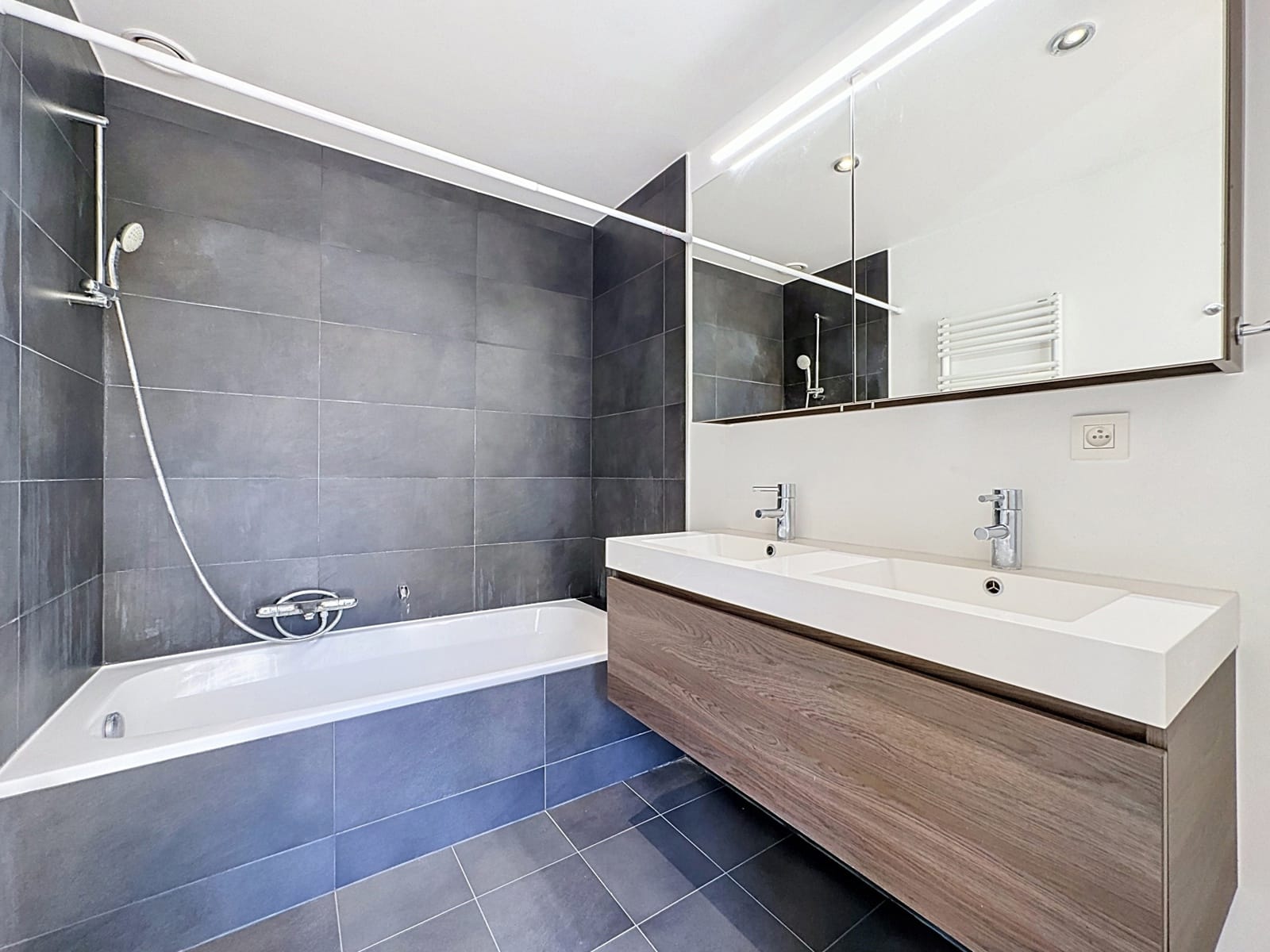 Moderne badkamer met dubbele wastafel – You Real Estate Agency Moderne badkamer in een appartement met ligbad, dubbele wastafel en houten badkamermeubel.