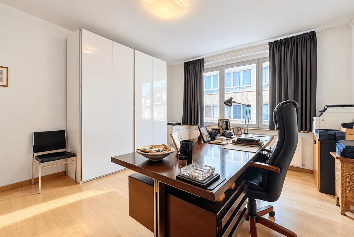 Modern home office with a wooden desk, black ergonomic chair, white storage cabinet, and a window with dark curtains outside the desk area.