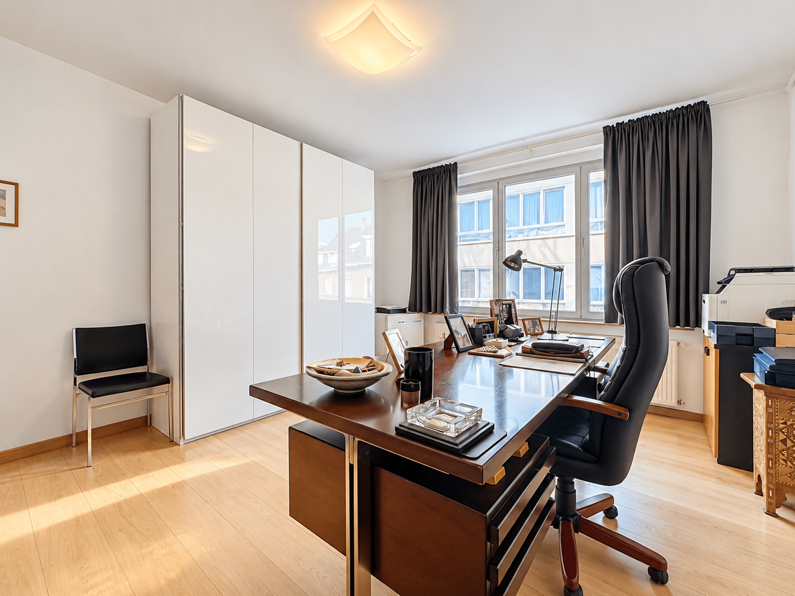 whise photo 10 Modern home office with a wooden desk, black ergonomic chair, white storage cabinet, and a window with dark curtains outside the desk area.