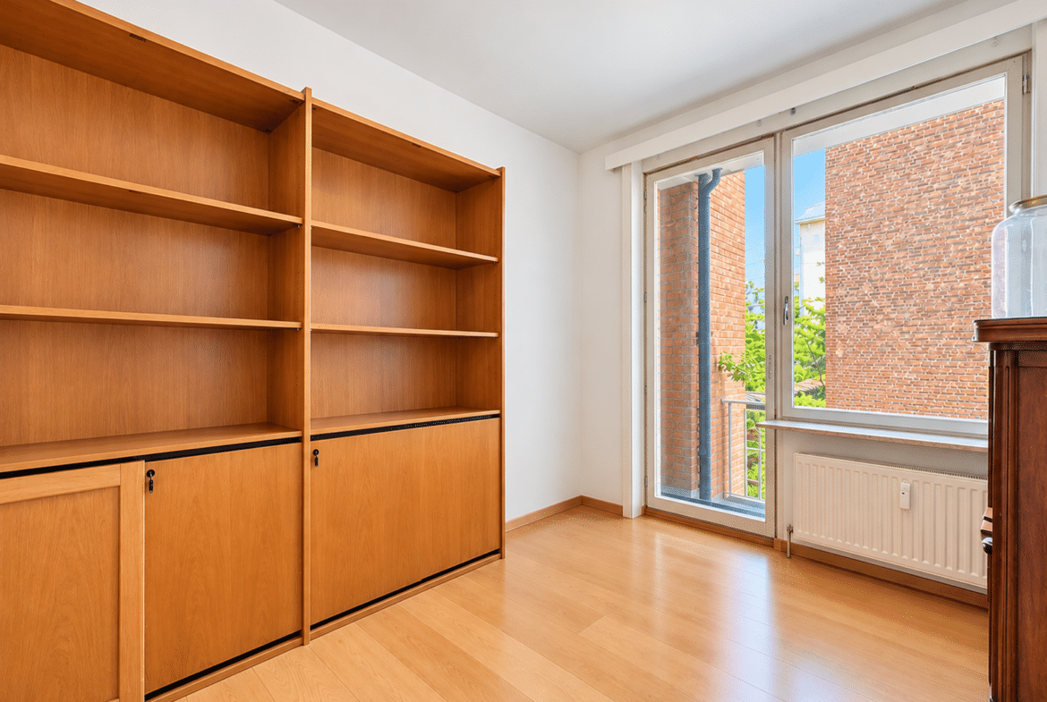 Room with tall wooden shelving unit along left wall, empty shelves, and a large window/door to a balcony on the right; brick building visible outside and a radiator beneath the window.