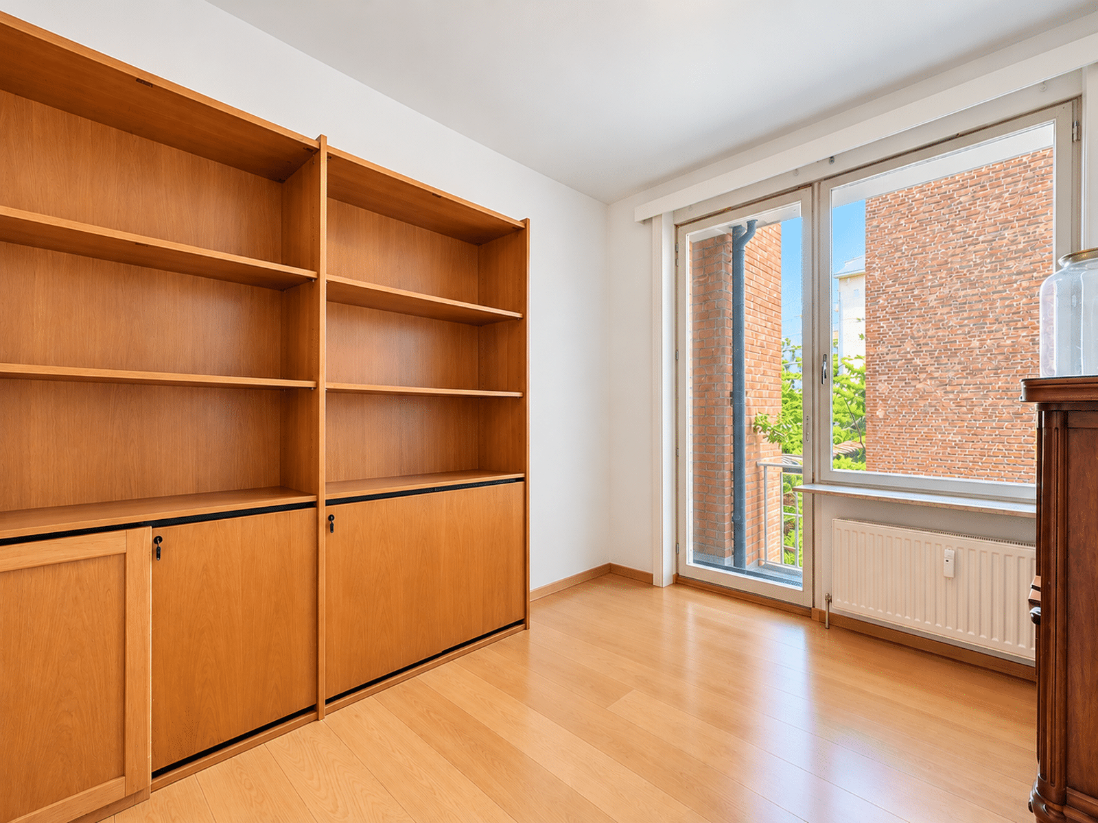 whise photo 15 Room with tall wooden shelving unit along left wall, empty shelves, and a large window/door to a balcony on the right; brick building visible outside and a radiator beneath the window.