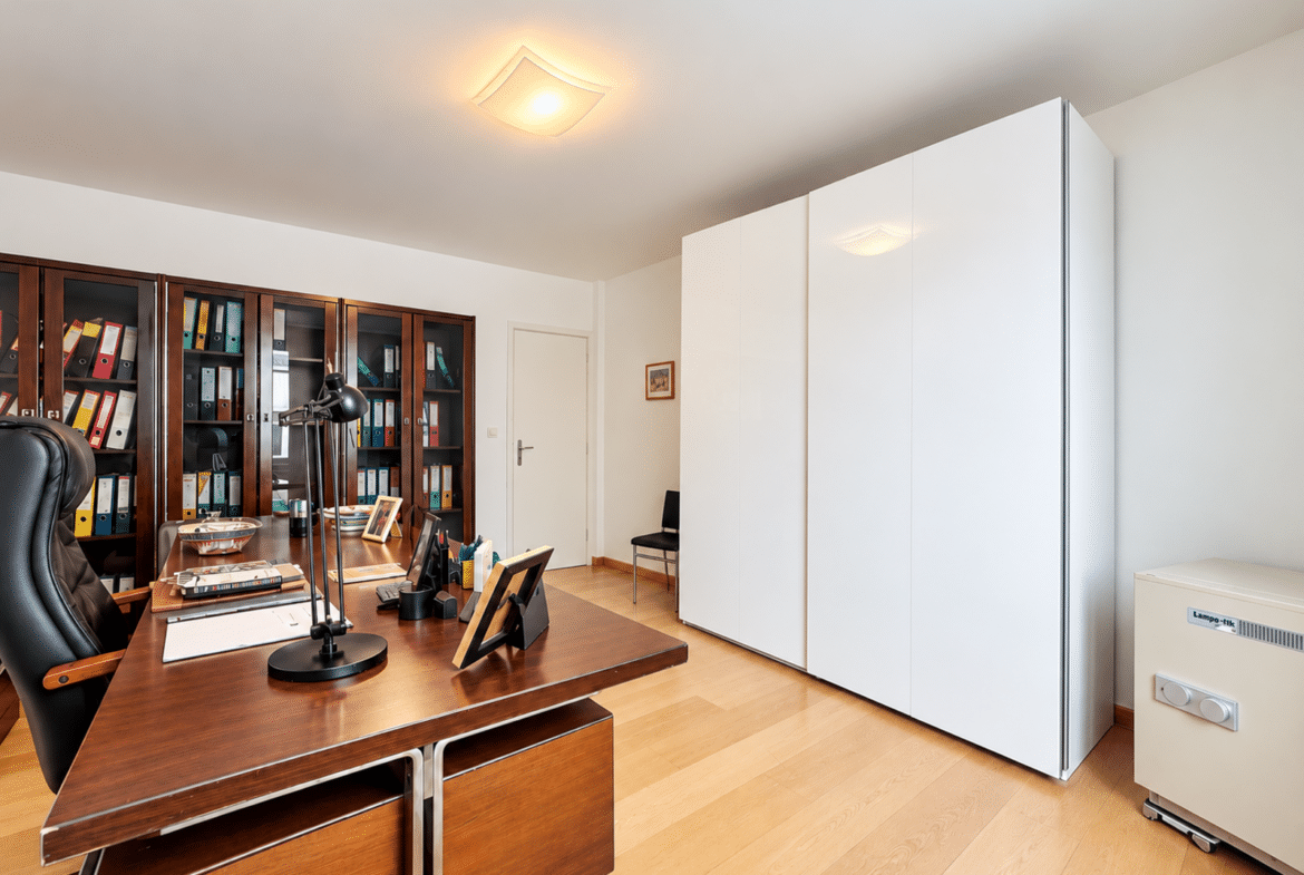 Office scene with a large wooden desk, black leather chair, desk lamp, and tall shelves filled with binders and folders behind it, plus white storage cabinets on the right.