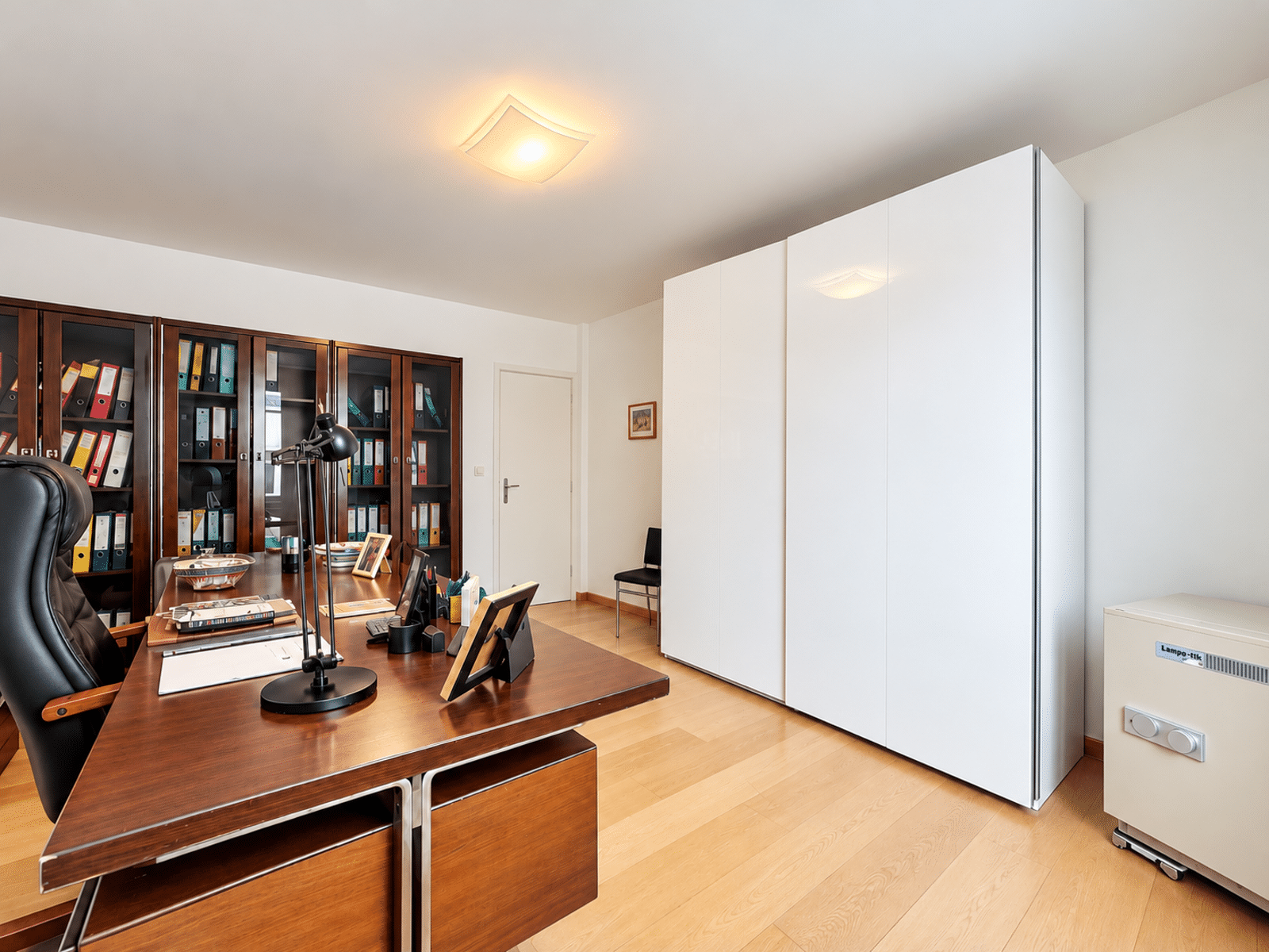 whise photo 12 Office scene with a large wooden desk, black leather chair, desk lamp, and tall shelves filled with binders and folders behind it, plus white storage cabinets on the right.