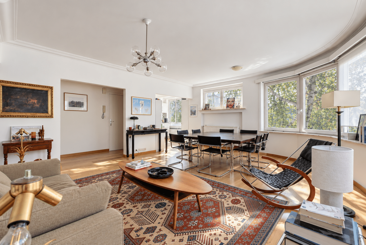 Bright, open living and dining room with a wooden dining table and black chairs by large windows letting in sunlight.